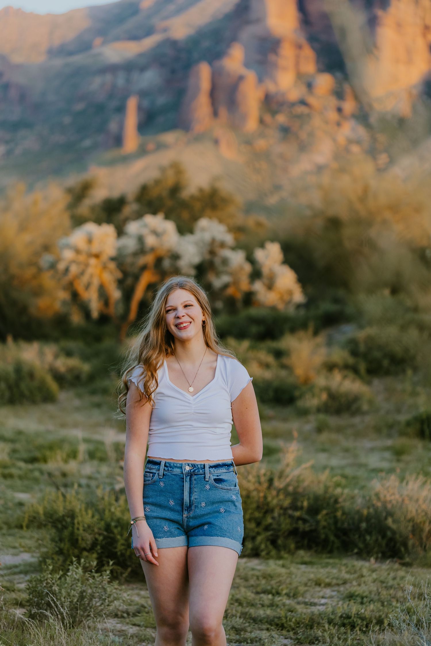 Woman in casual attire poses in desert landscape during golden hour with mountains behind.