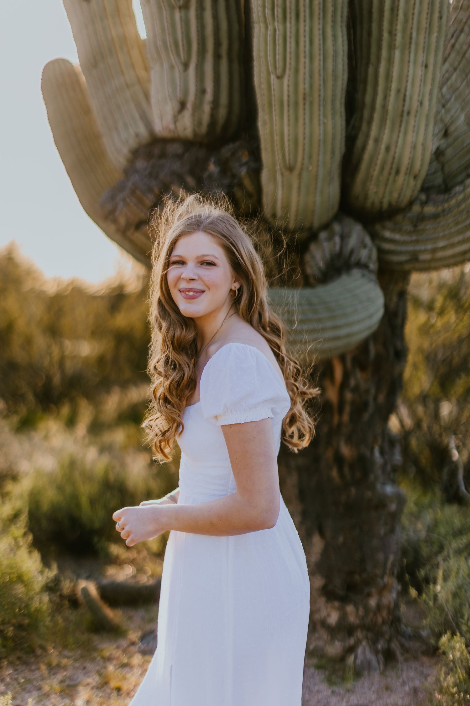 Portrait in a flowing white dress against desert landscape with saguaro cactus at sunset.