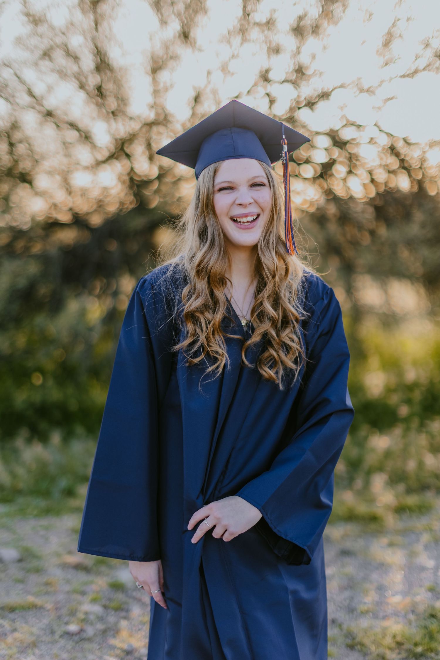 Graduate in navy blue cap and gown poses outdoors during golden hour celebration photoshoot.
