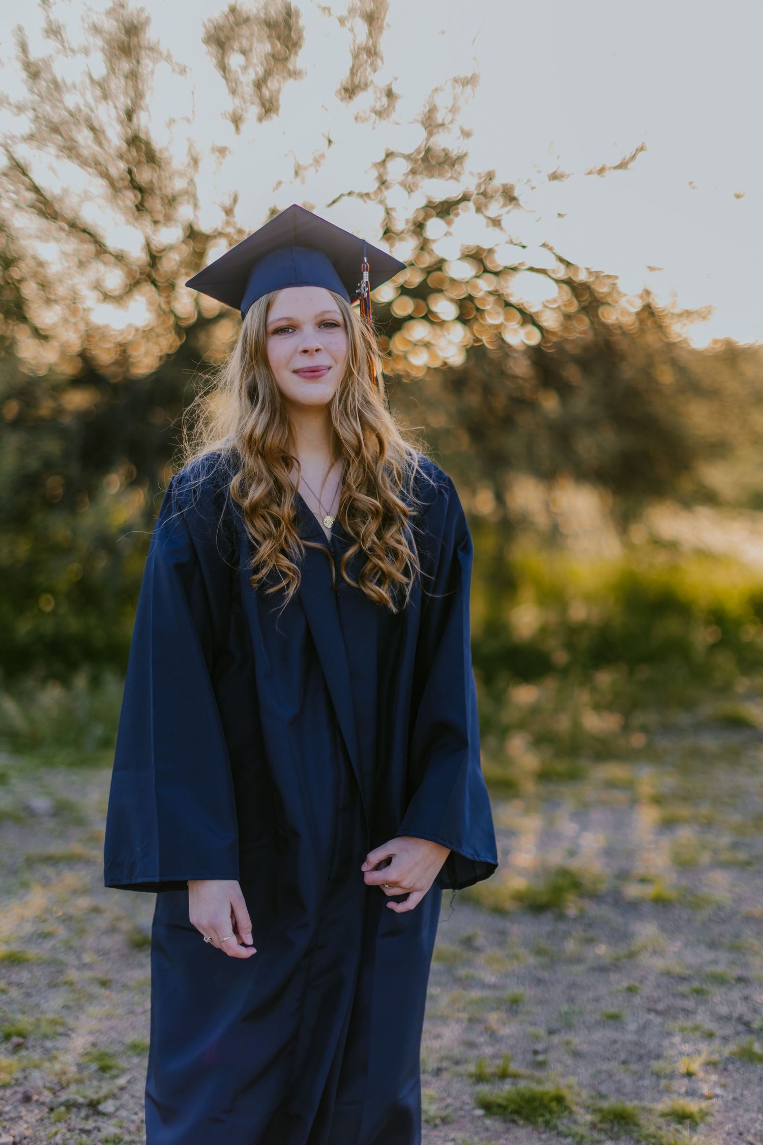 Graduate stands in traditional navy graduation attire against natural outdoor backdrop at sunset.