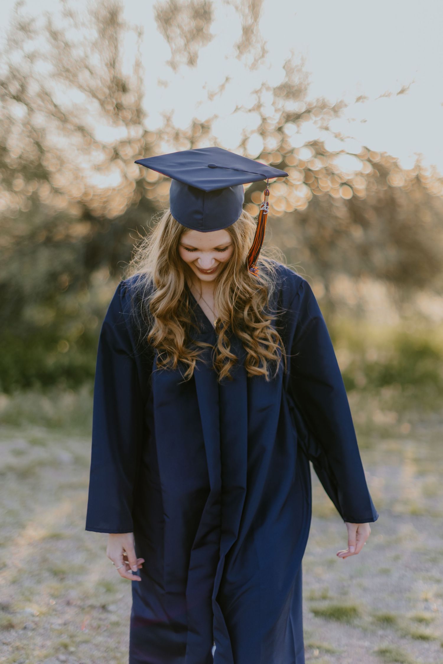 Graduate in navy academic regalia looks down with joy during outdoor graduation photos.