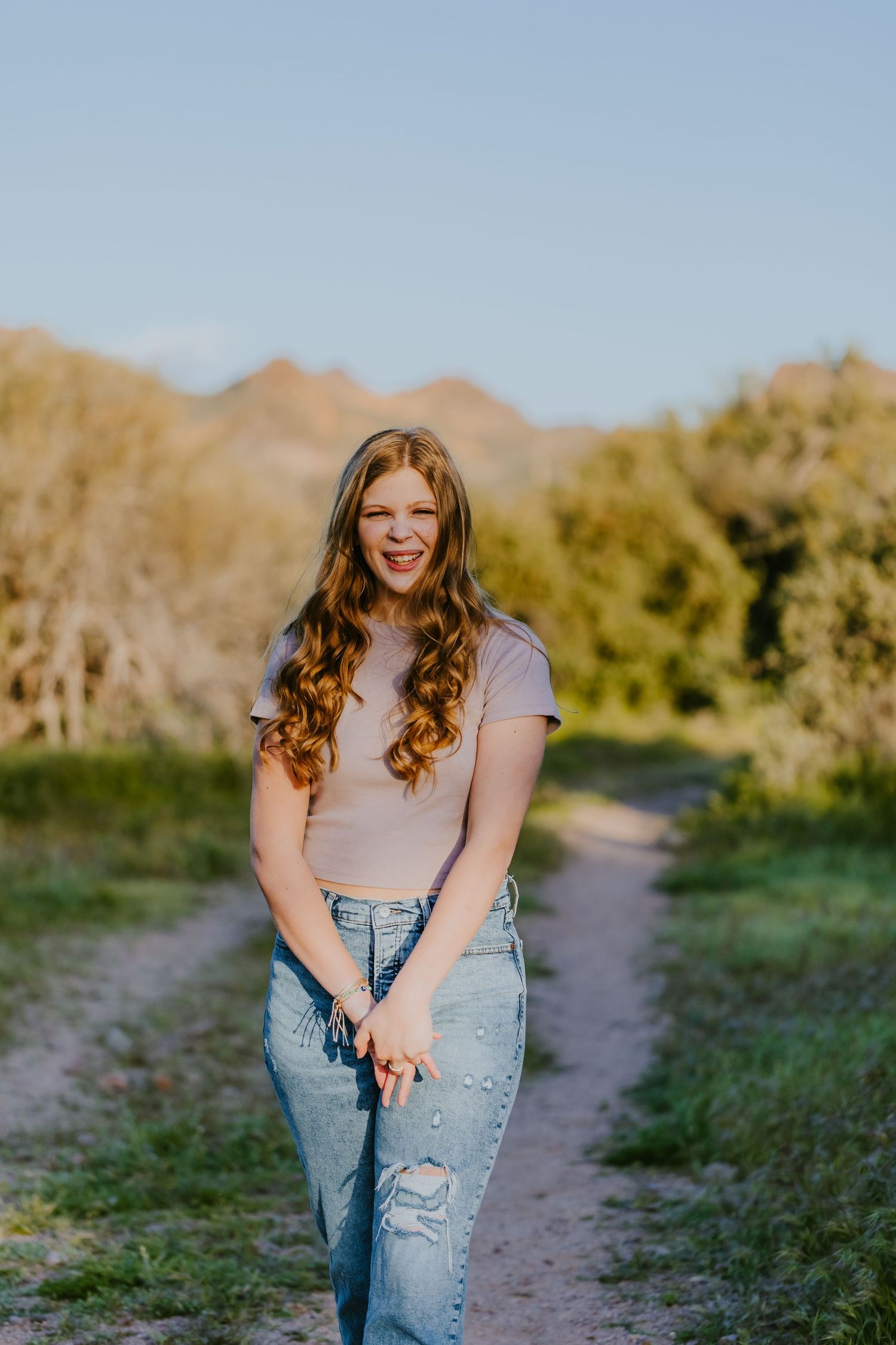 A casual outdoor portrait showing long curly hair, beige crop top and distressed jeans against a mountain backdrop.