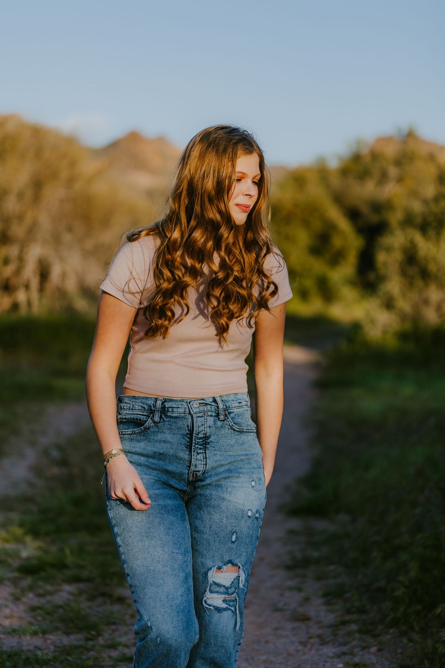 Natural outdoor photo featuring wavy brown hair, neutral top and ripped jeans on a dirt path.