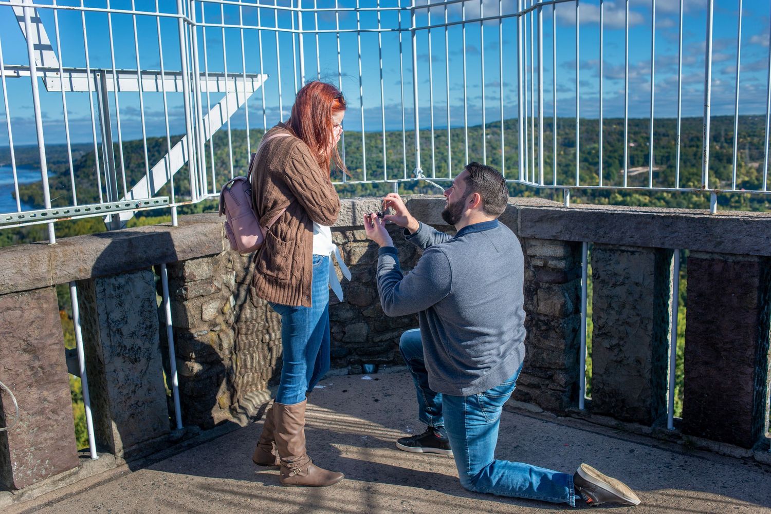 A romantic proposal taking place at a scenic mountain overlook on a bright sunny day with blue skies in the background.
