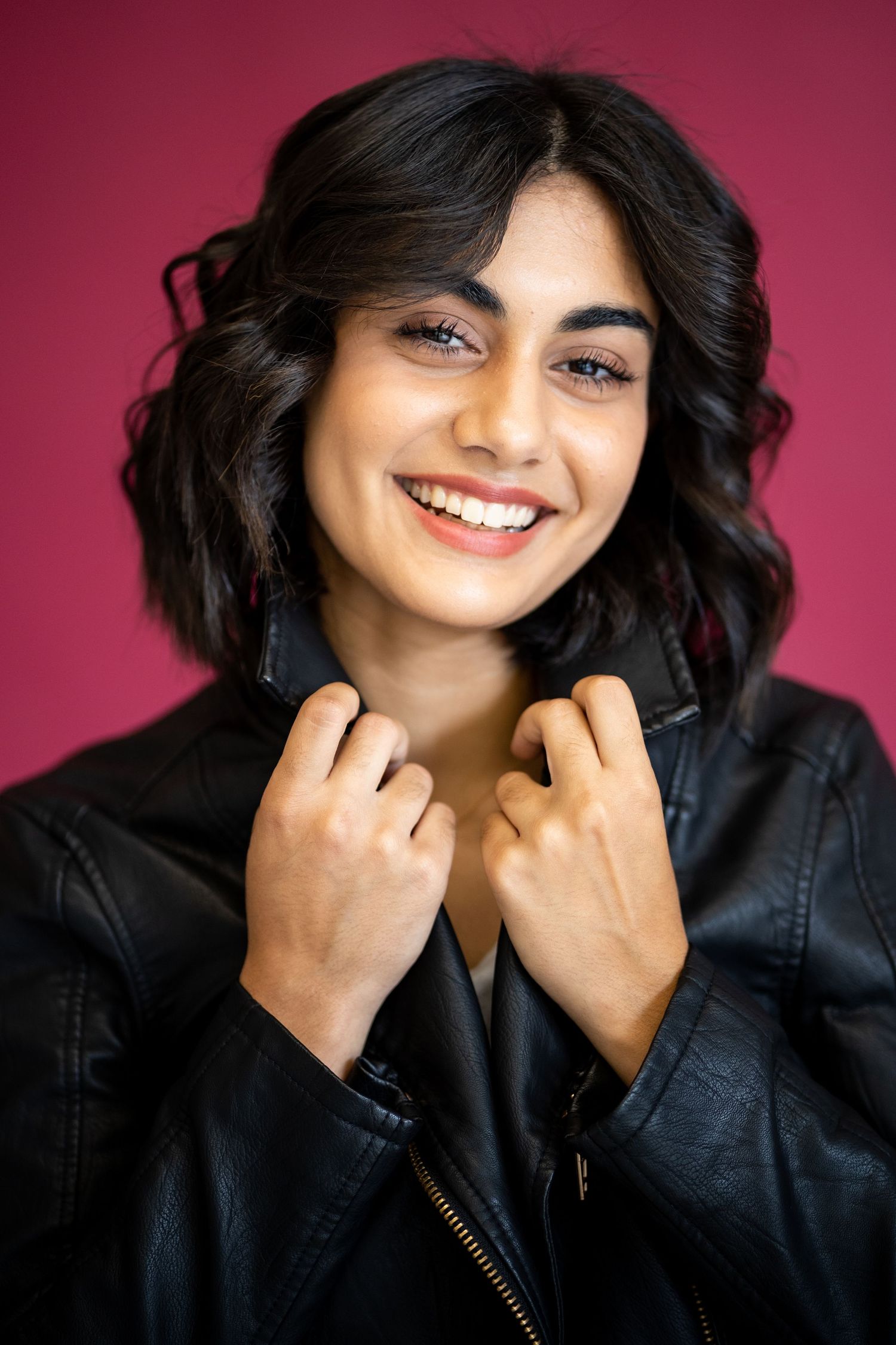 A high school senior in black leather jacket smiling against burgundy background during studio photoshoot.