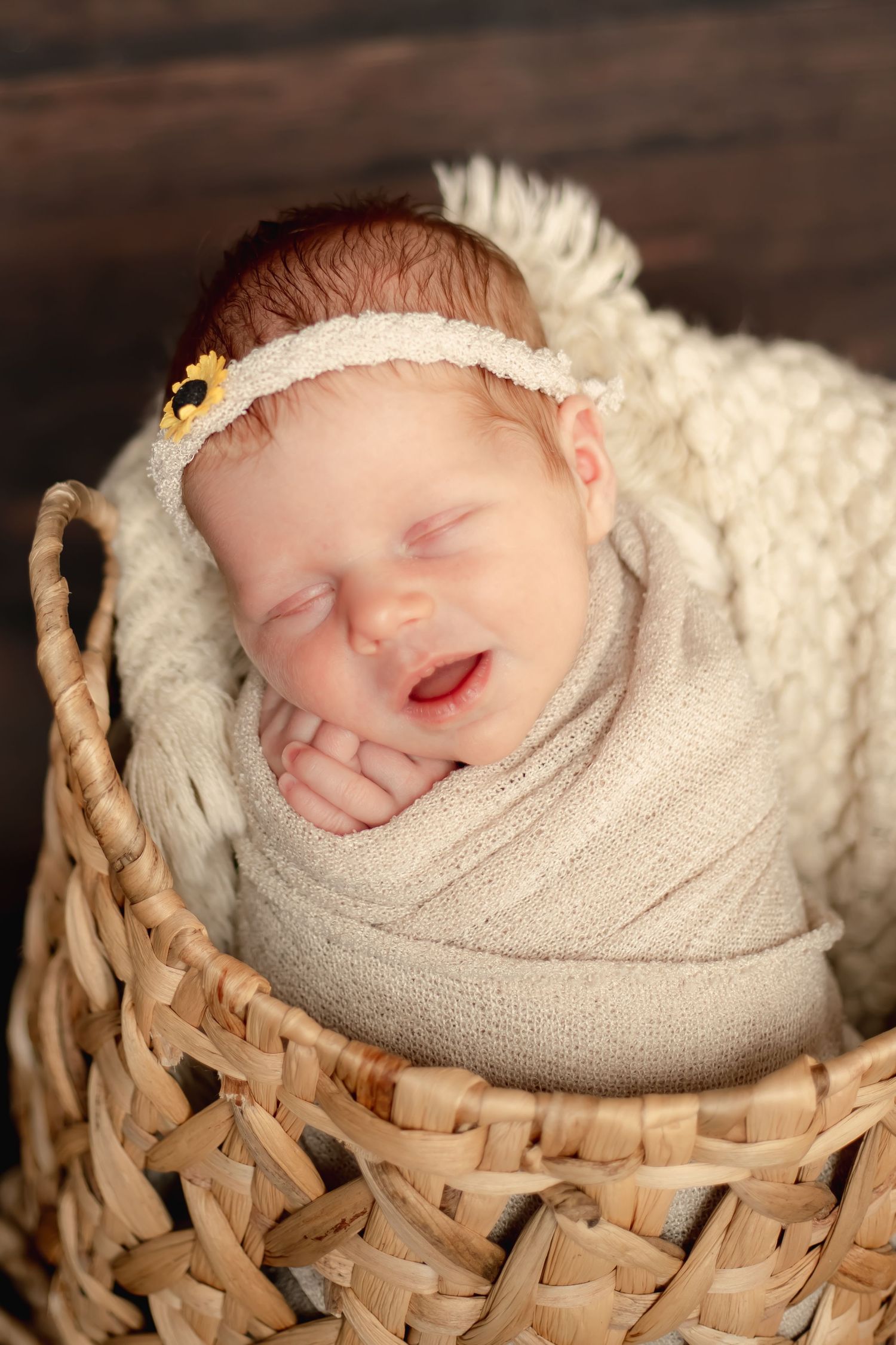 Smiling newborn in woven basket wearing delicate headband wrapped in cream fabric.