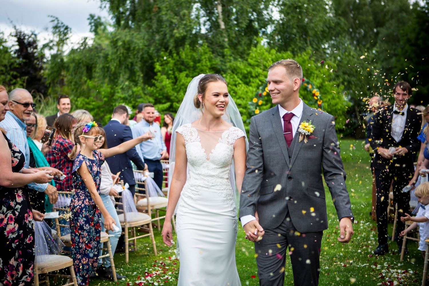 Newlyweds walk down outdoor aisle as guests throw confetti at summer garden wedding.