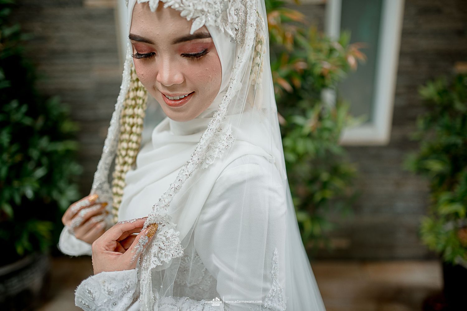 A bride in white hijab and wedding dress smiles while touching her traditional bridal accessories.