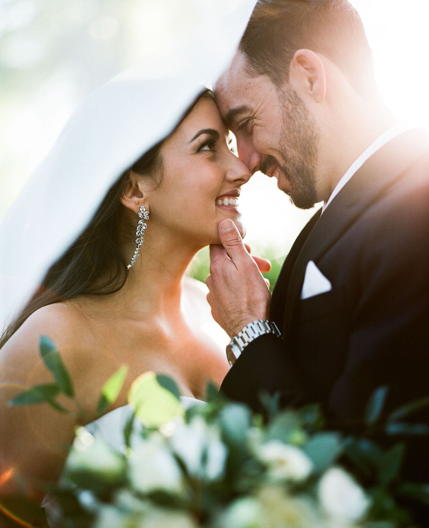 Intimate wedding portrait captured in soft golden sunlight with greenery in background.