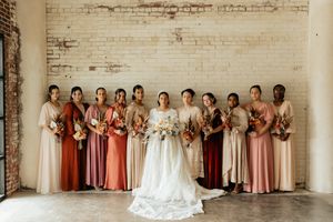 Wedding party in coordinating pink and mauve dresses poses against a white brick wall.