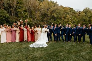 Large wedding party in formal attire poses outdoors against an autumn landscape.