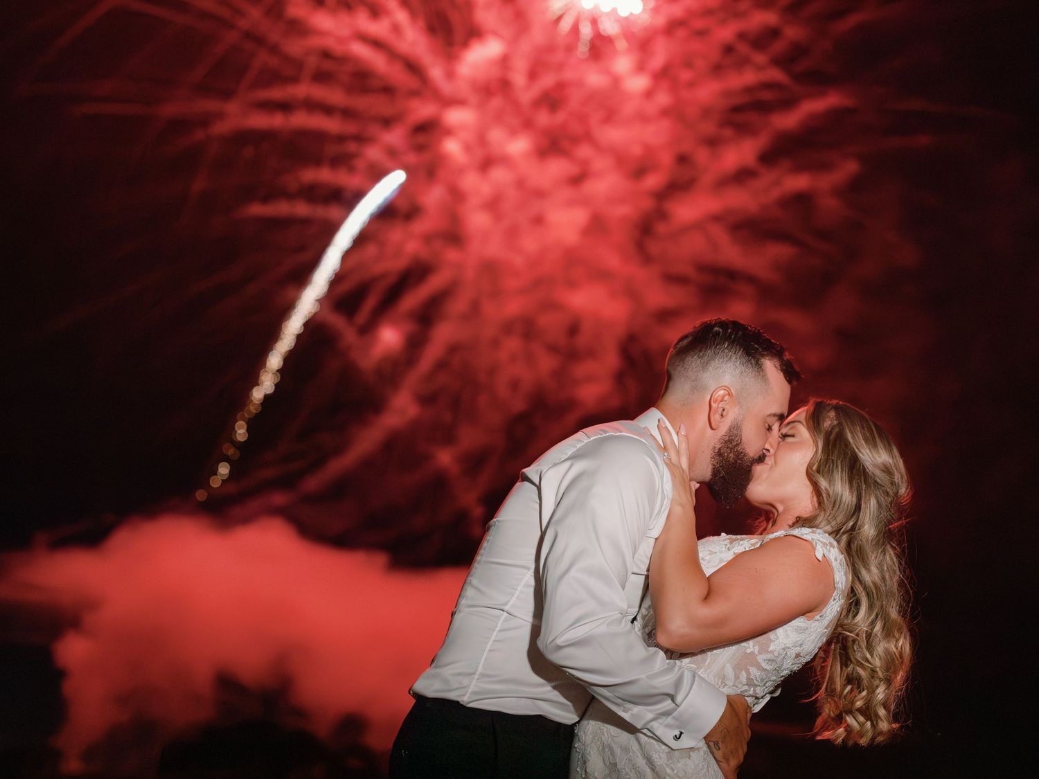 The groom dips his bride into a kiss, a bright, red firework exploding in the background.
