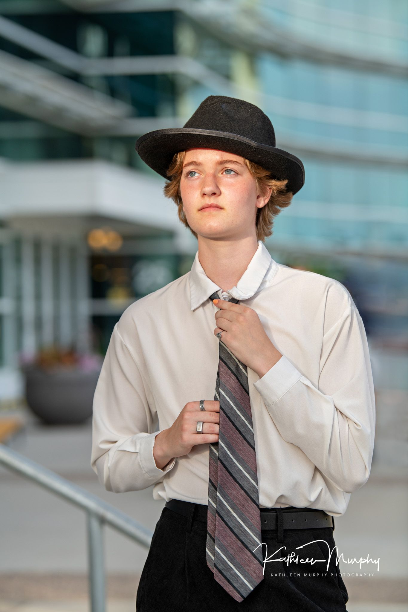 Professional portrait wearing hat and striped tie against modern glass building exterior.
