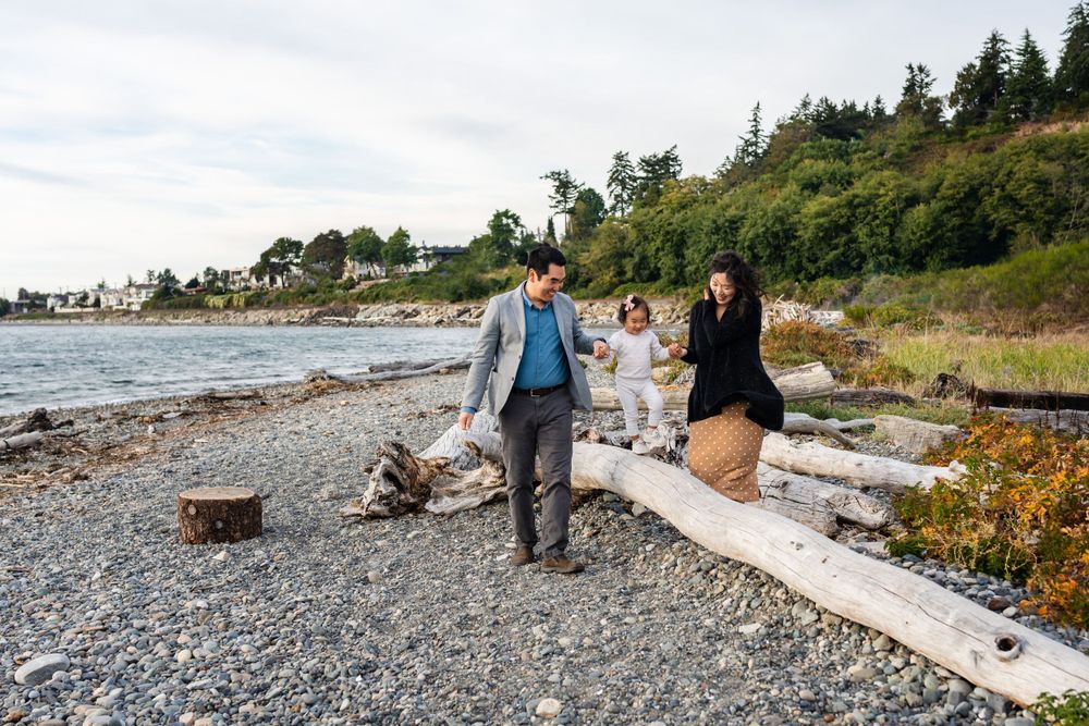 Parents holding hands with their daughter and walking on driftwood at Richmond Beach Saltwater Park.