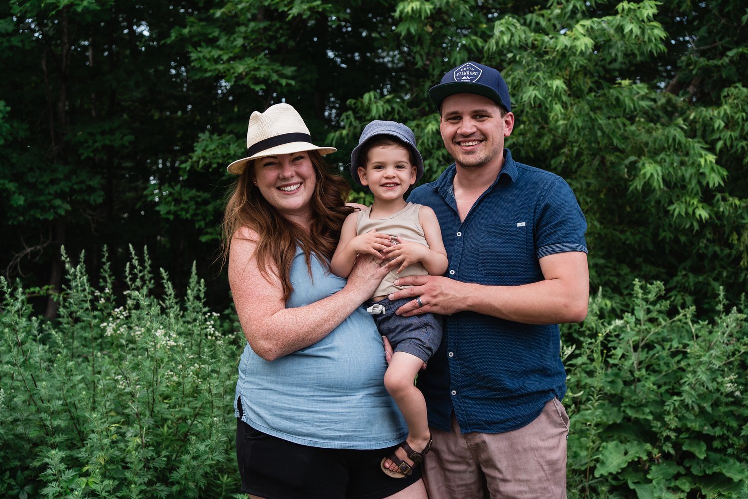 Family of Mother, Father and toddler son smile for the camera in front of a lush green forest