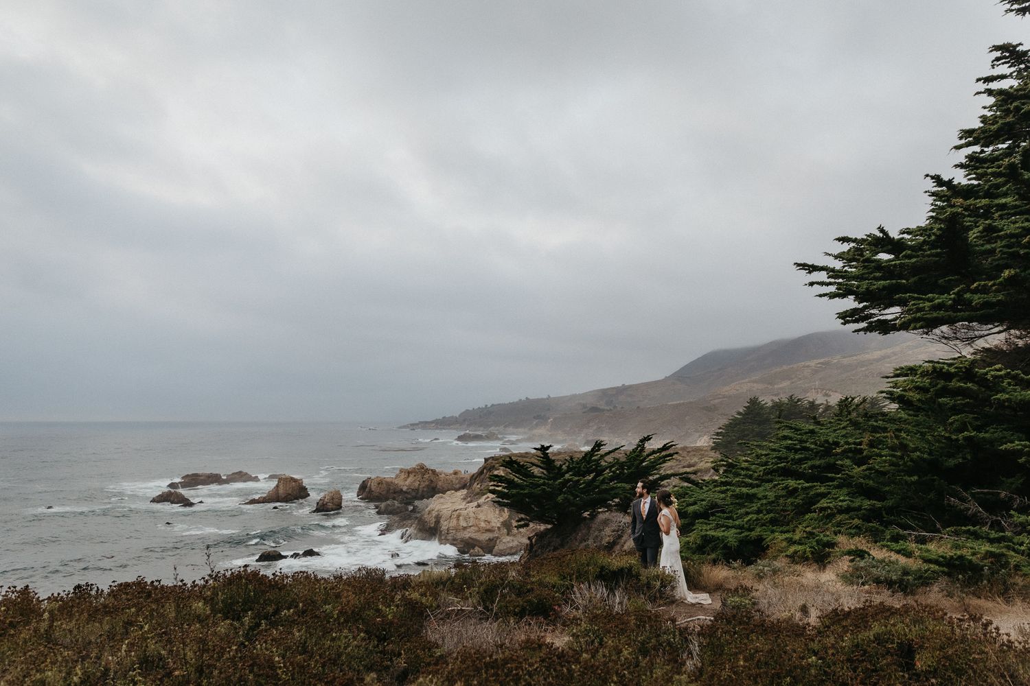 Panoramic coastal wedding photo with rugged cliffs and moody skies.