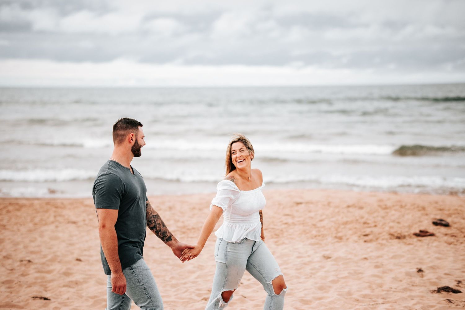 A couple holds hands and laughs together while walking on a scenic beach under cloudy skies.