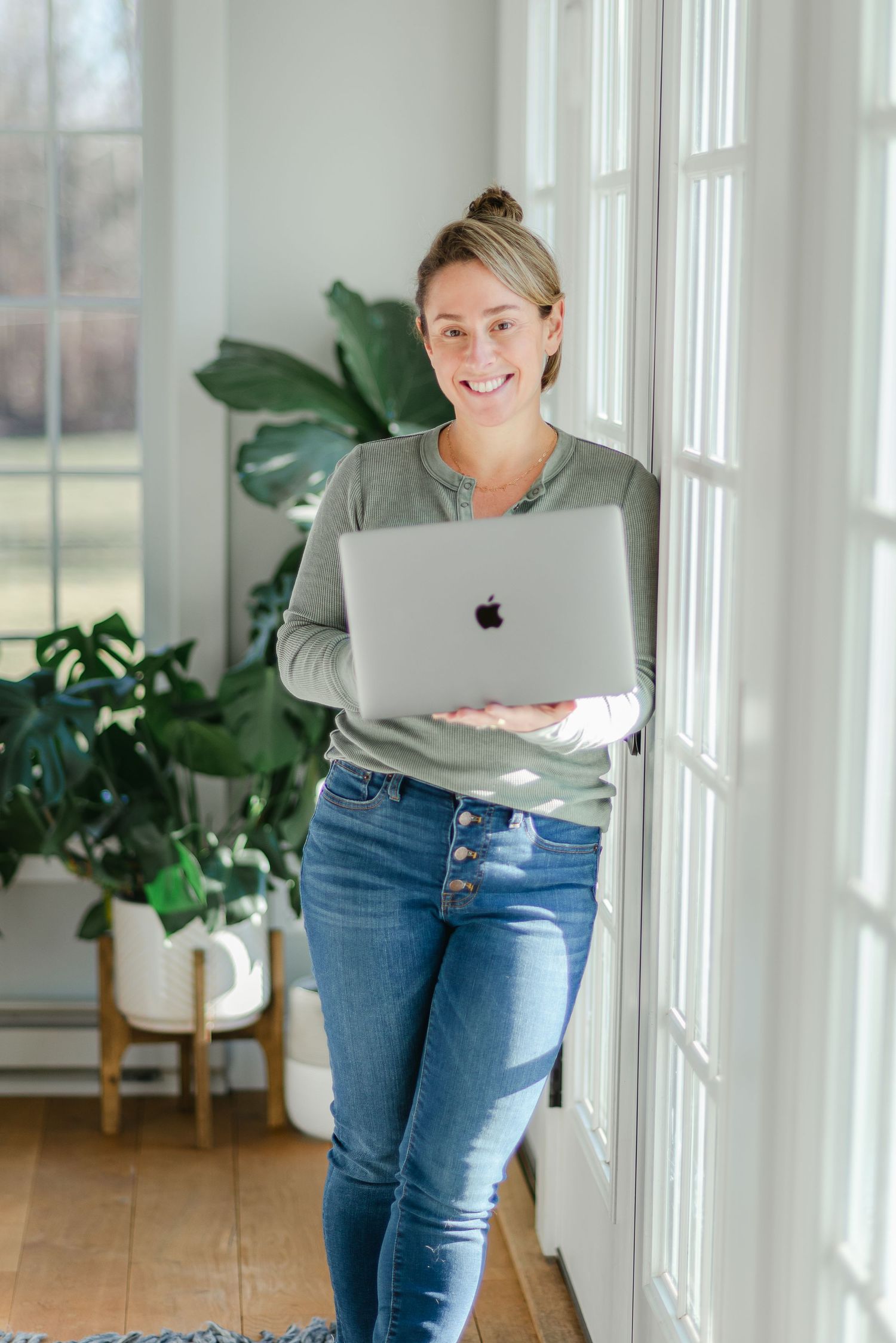 Professional working woman holds MacBook laptop while standing by bright window with indoor plants.