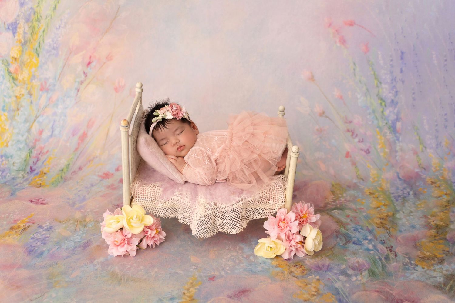 A tiny baby sleeps peacefully in a decorative bed with pink floral accents and pastel backdrop.