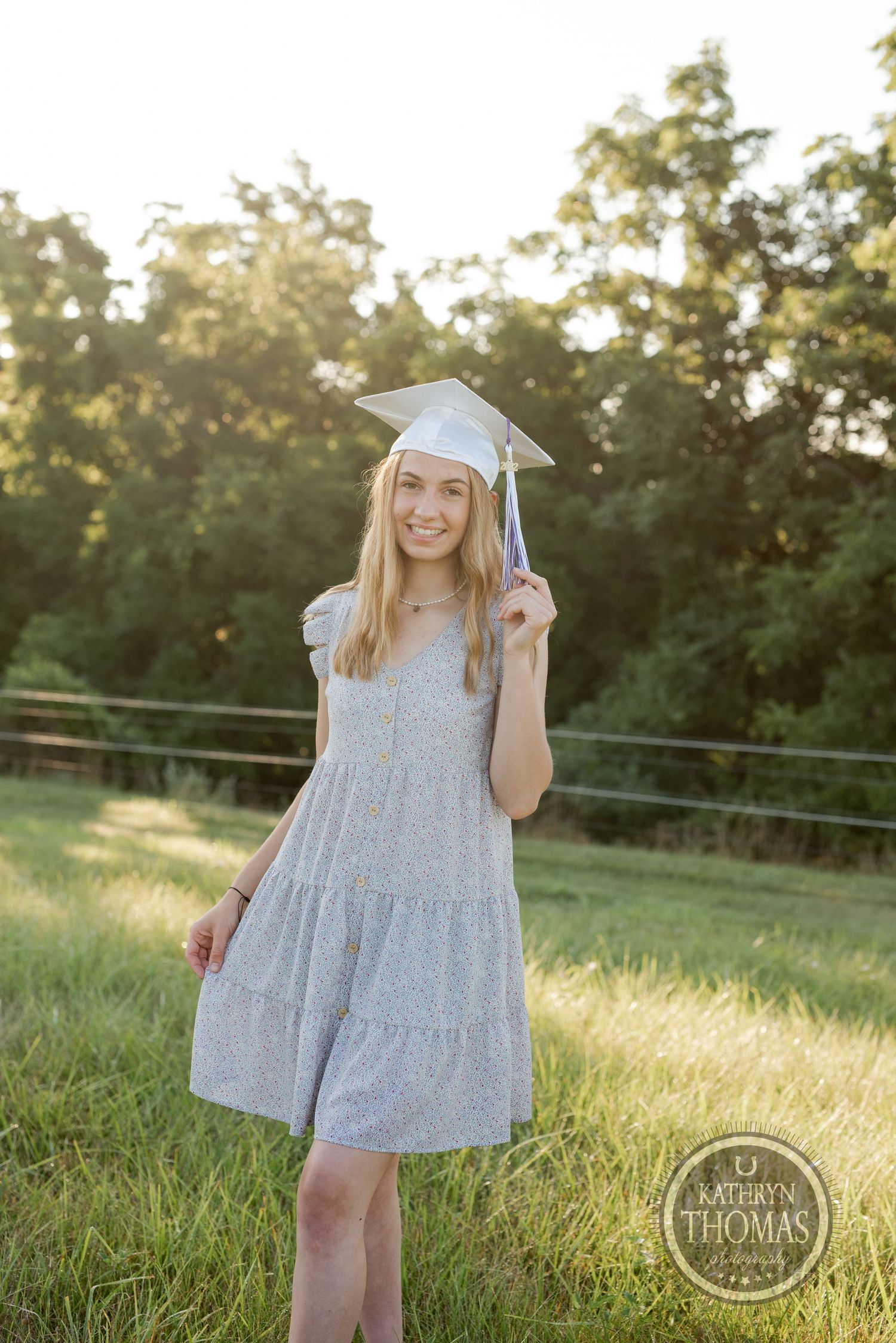 A graduate poses in a light floral dress and cap against a sunlit rural background.