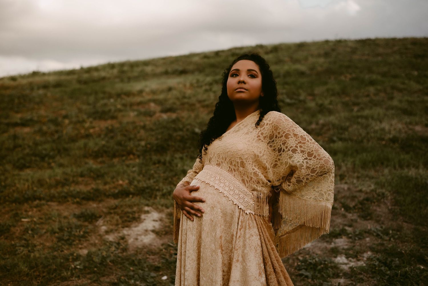 Dramatic maternity photo featuring flowing beige lace dress against overcast sky on hilltop.