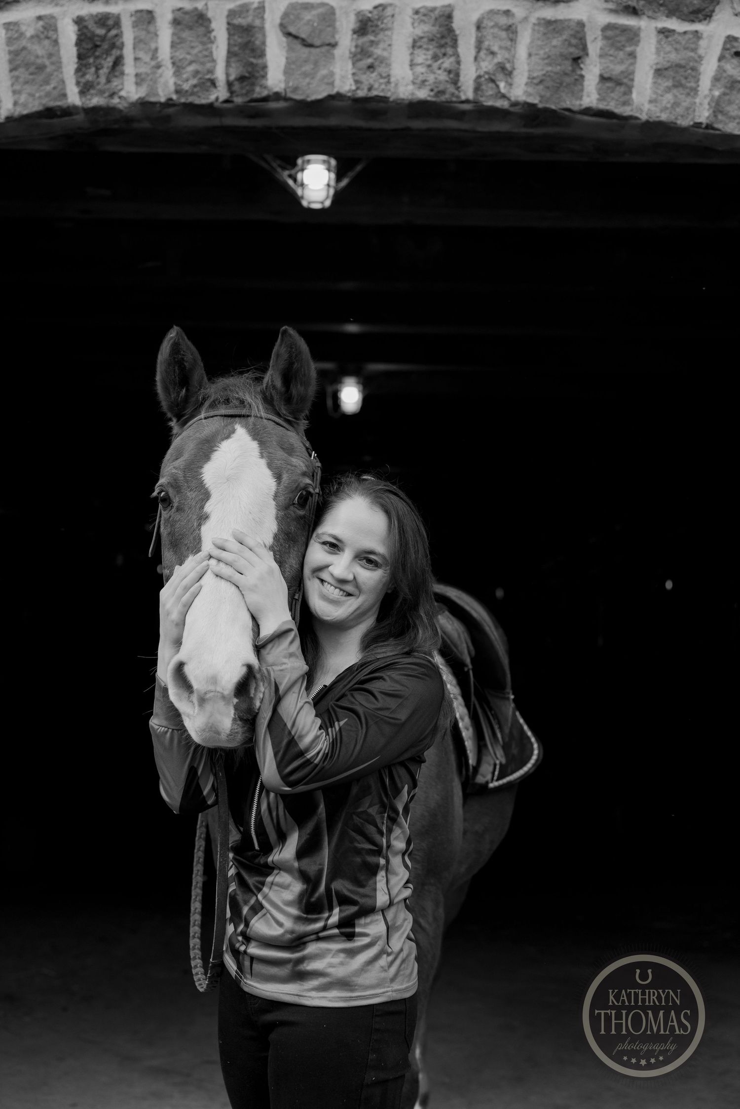 A black and white portrait of a horse and rider standing in a rustic barn doorway.
