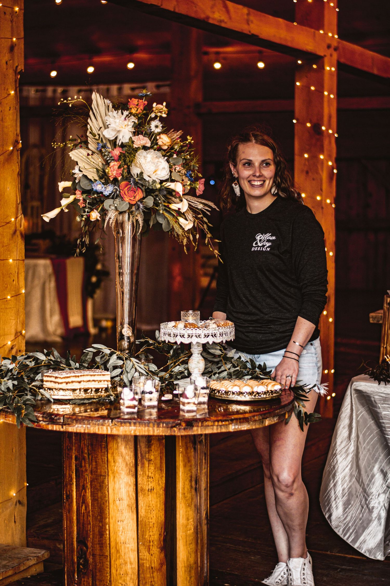A dessert display table features a tall floral arrangement and tiered cake stands in a rustic venue.