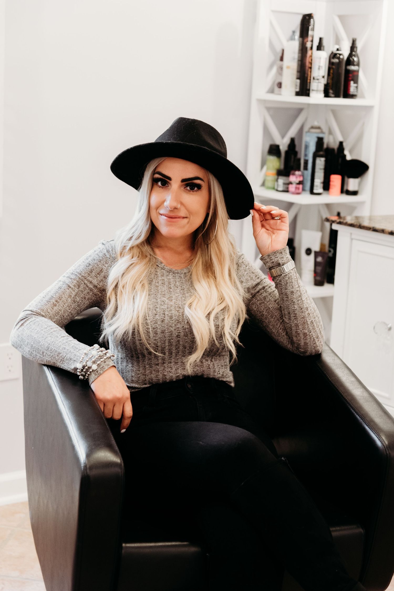 Stylish woman in gray sweater and black hat poses in salon chair with shelving in background.