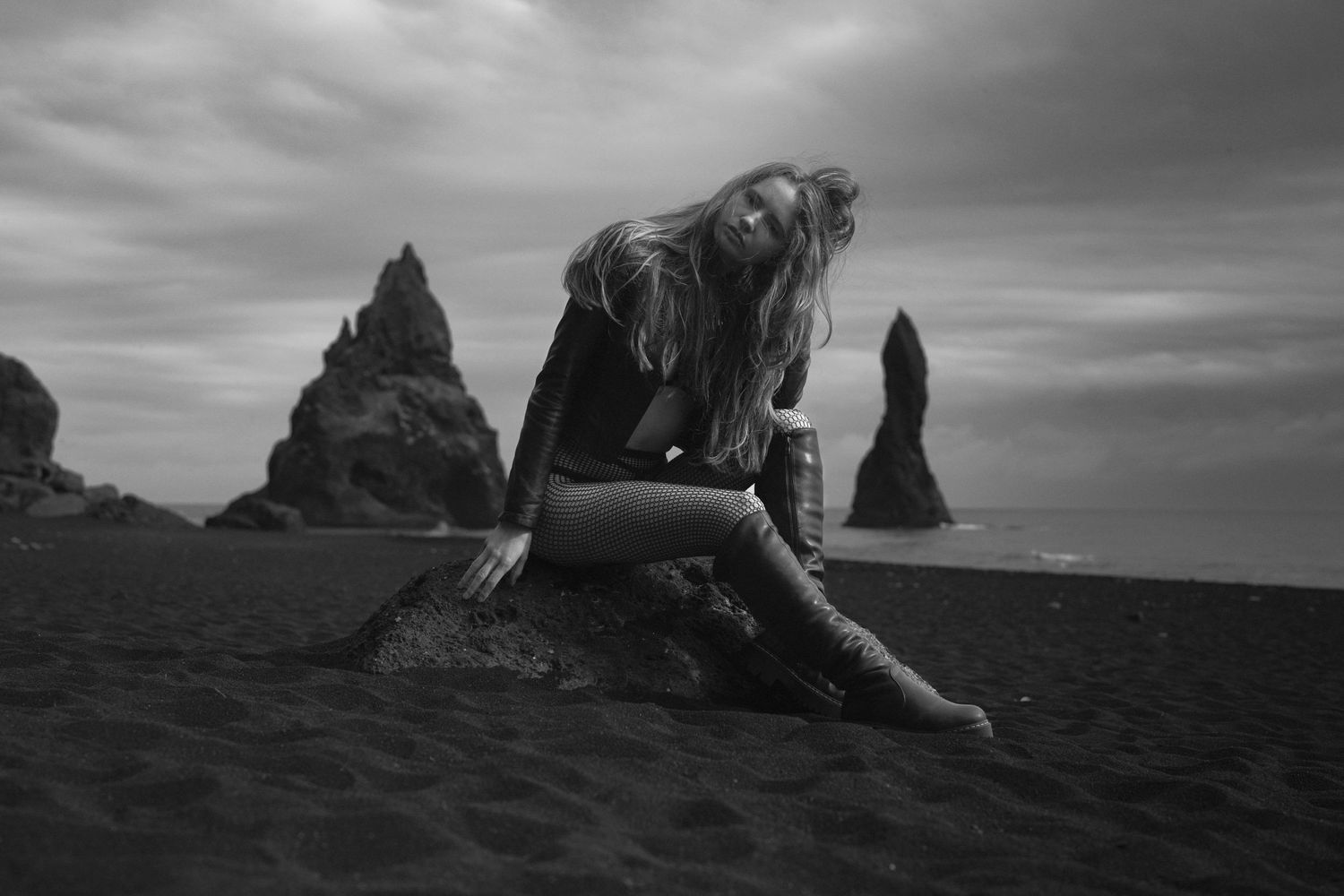 An artistic black and white photo of a figure posed dramatically on a beach with rock formations.