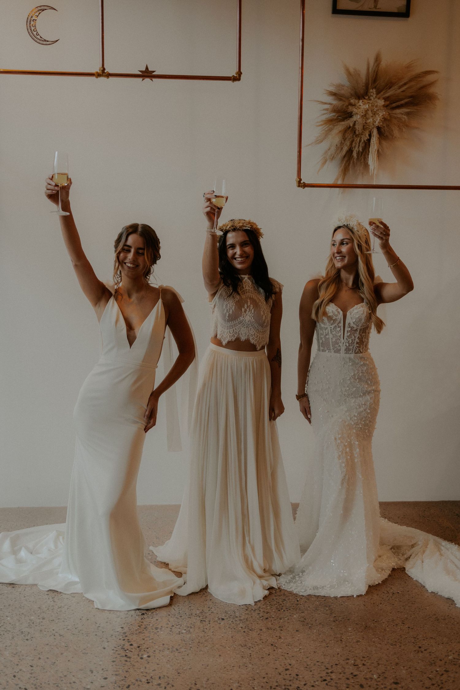 Three brides in white dresses raise champagne glasses in celebration against a neutral wall with moon decor.