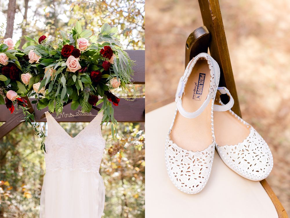 A delicate lace wedding dress hangs beside white floral shoes and pink flower arrangements.