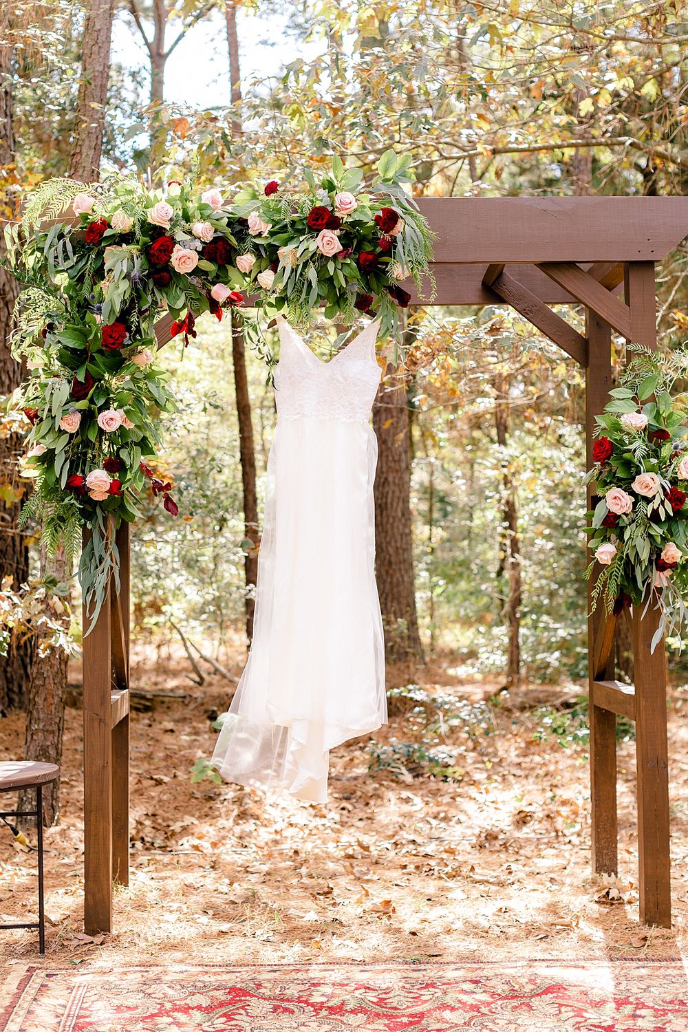 White wedding dress hanging on wooden arch decorated with lush red and green floral arrangements.