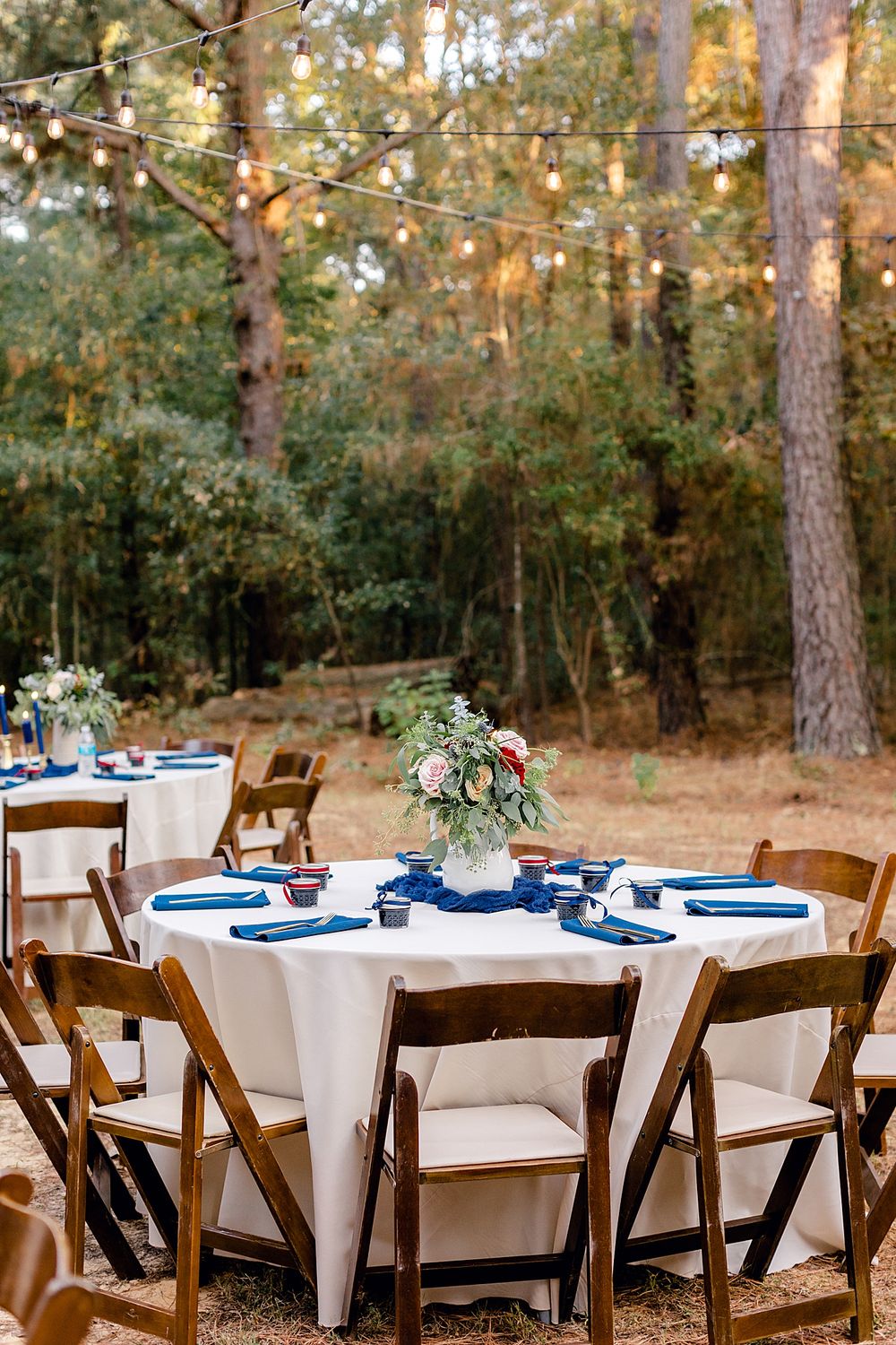 Outdoor wedding reception table with white linens, navy accents, and wooden chairs under string lights.