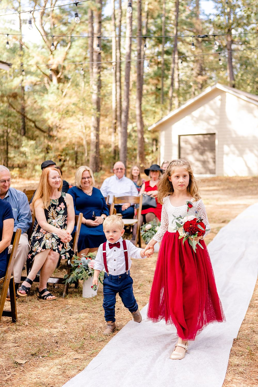 An outdoor wedding ceremony with a flower girl in red dress walking down a white aisle runner.