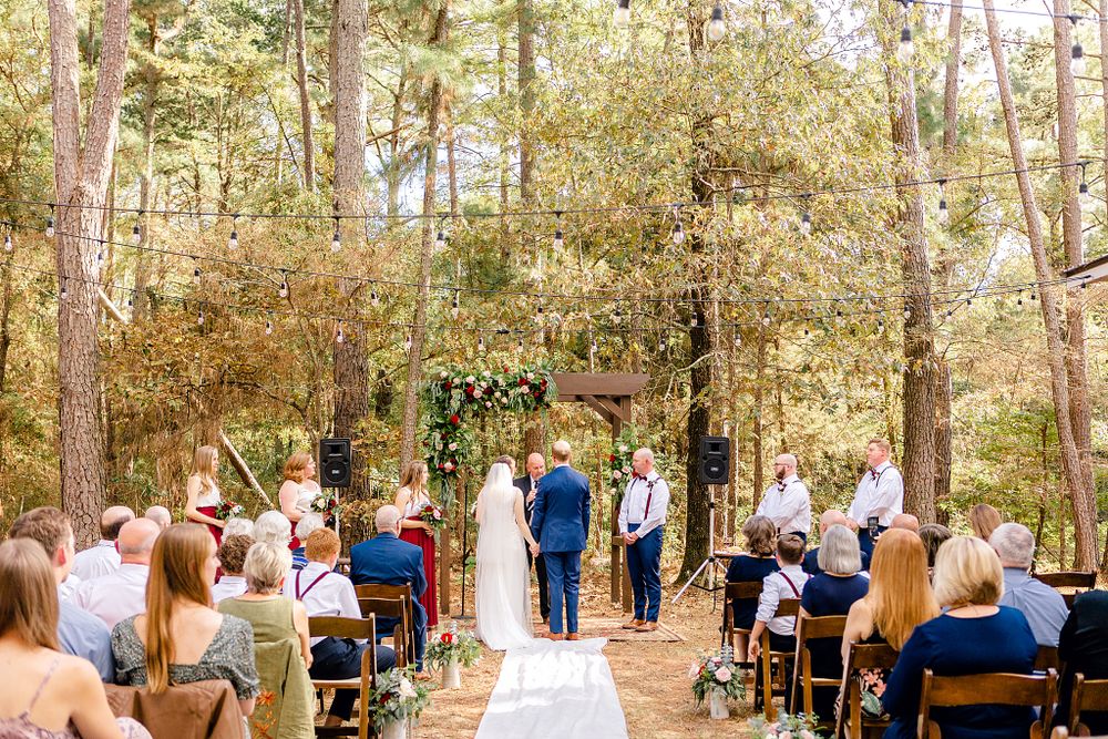 Outdoor forest wedding ceremony with guests seated on white chairs and natural woodland backdrop.