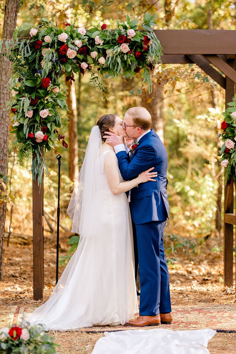 Couple sharing first kiss under floral arch decorated with red roses and greenery.