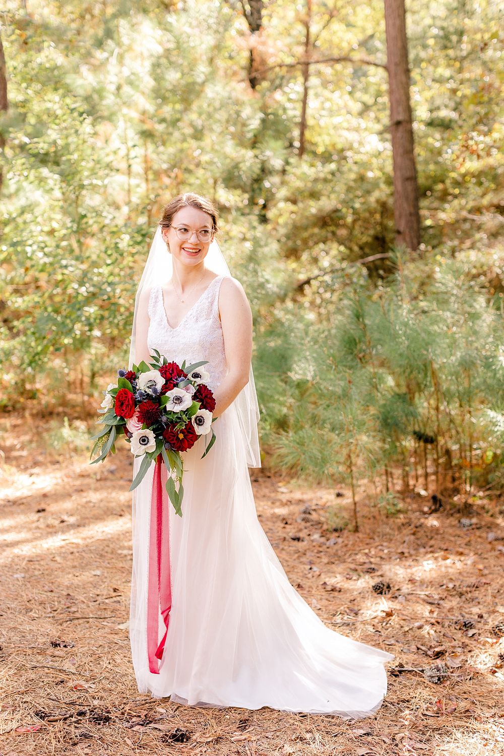 Beautiful wedding portrait of bride holding red and white bouquet in sunlit forest.