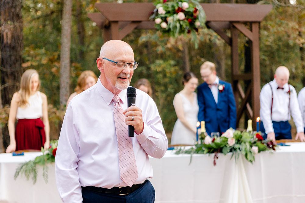 A speaker gives a toast at an outdoor wedding reception under a wooden pergola.