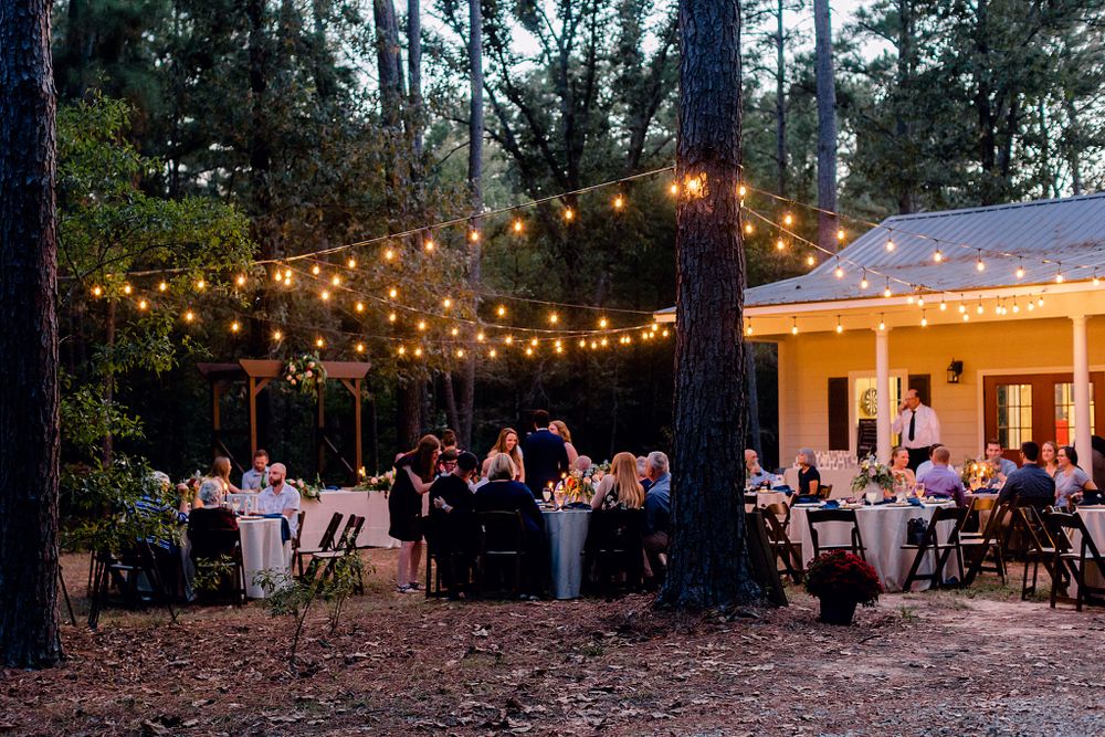 An evening outdoor wedding reception with string lights illuminating tables under tall pine trees.