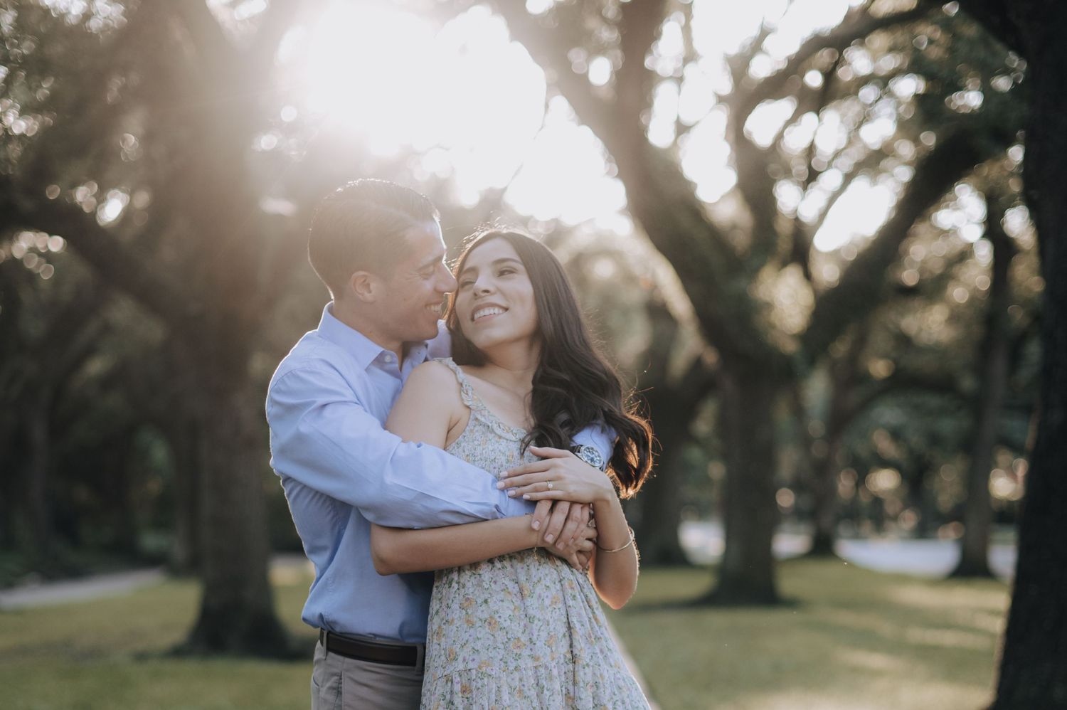 A romantic couple embraces in a sunlit park with trees in the background.
