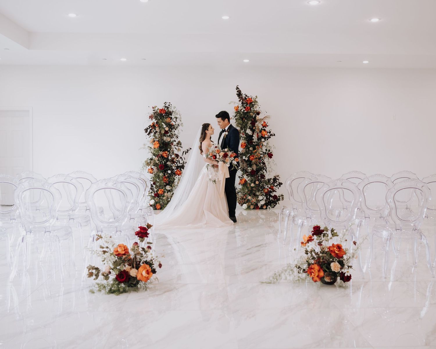 Elegant white wedding ceremony setup with floral arch and ghost chairs in modern minimalist space.