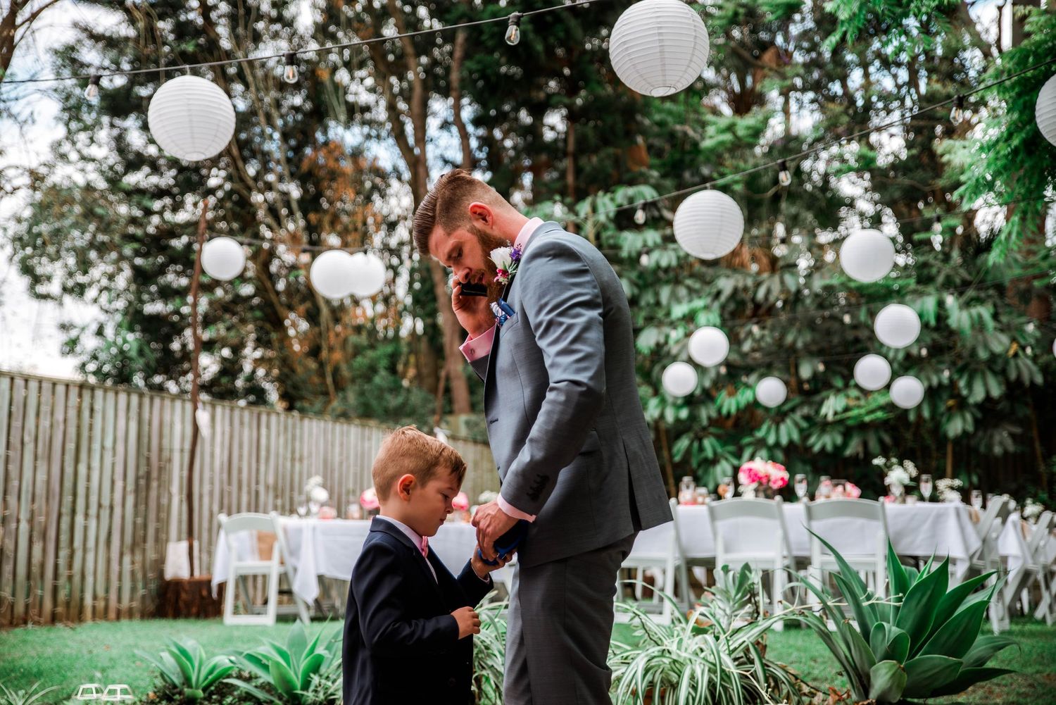 A tender moment between a groom and young boy at an outdoor garden wedding reception with white lanterns.