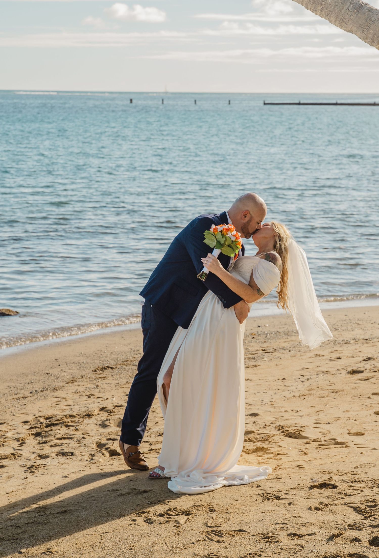 Newlyweds share a romantic kiss on a sandy beach with ocean backdrop.