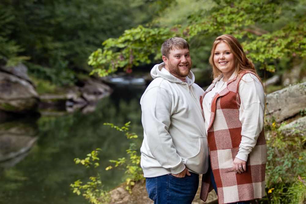 A couple poses together near a serene stream in an autumn forest setting.
