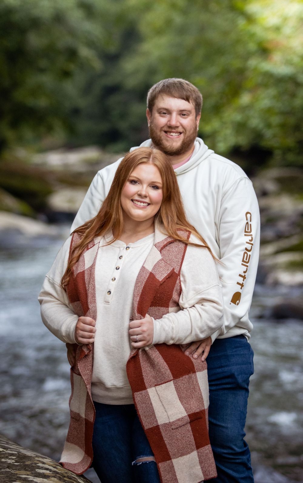 A romantic outdoor portrait by a flowing stream with fall foliage in the background.