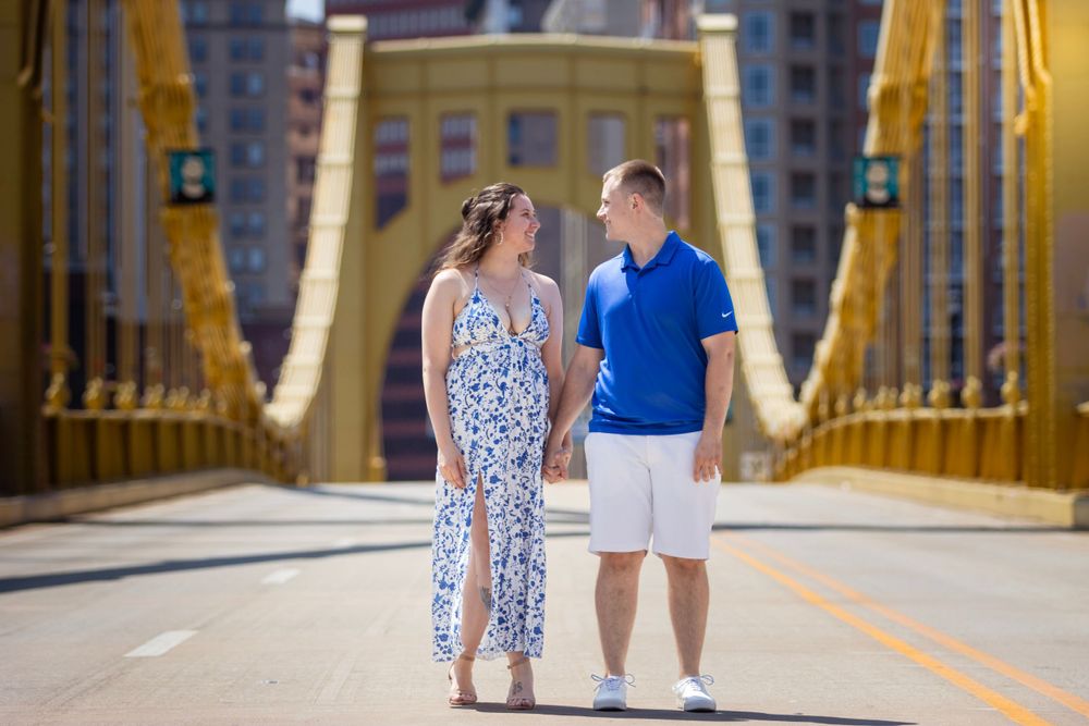 A couple standing together on Pittsburgh's iconic yellow Roberto Clemente Bridge on a sunny day.