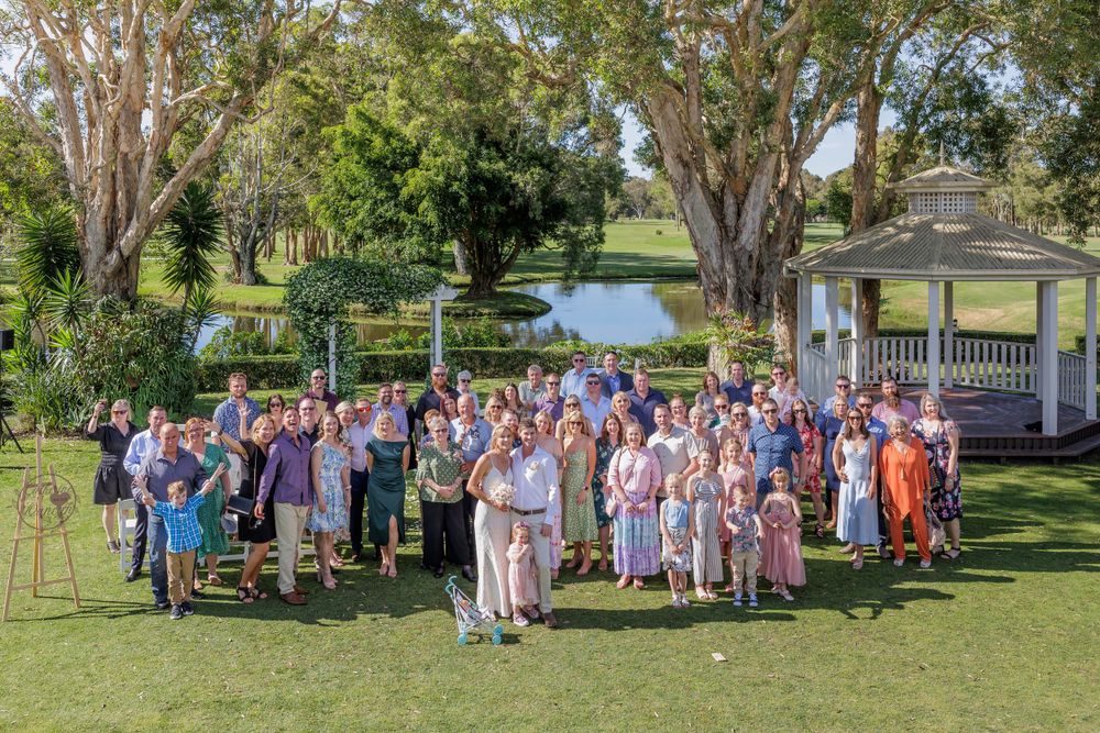 Large group wedding photo with guests gathered under trees in a park setting.