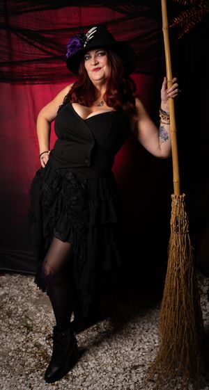 Dark-themed witch photoshoot featuring a figure in black holding a broomstick against a red backdrop.