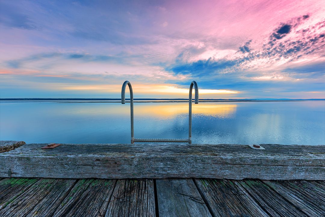 Swimming pool ladder silhouetted against a pastel sunset over calm ocean waters.