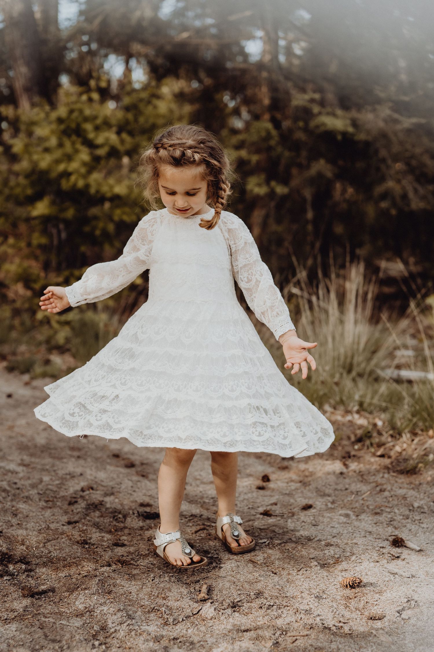A young child in a white lace dress twirls playfully on a dirt path outdoors.